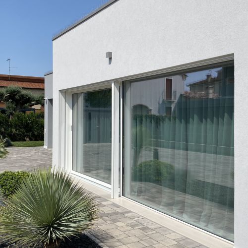 Facciata di una casa moderna di nuova costruzione con alzante scorrevole protetto da tende motorizzate integrate colr grigio. Serramenti in legno-alluminio bianchi integrati nel design minimalista dell'abitazione.