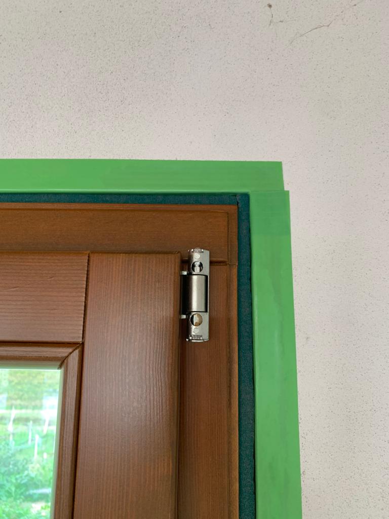 Dettaglio ravvicinato della cerniera di una finestra in legno posata a regola d'arte, con profilo verde di protezione ancora visibile.