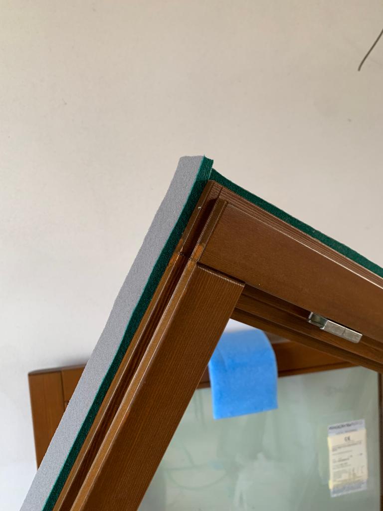 Dettaglio del nastro autoespandente grigio applicato sull'angolo esterno del telaio della finestra in legno.